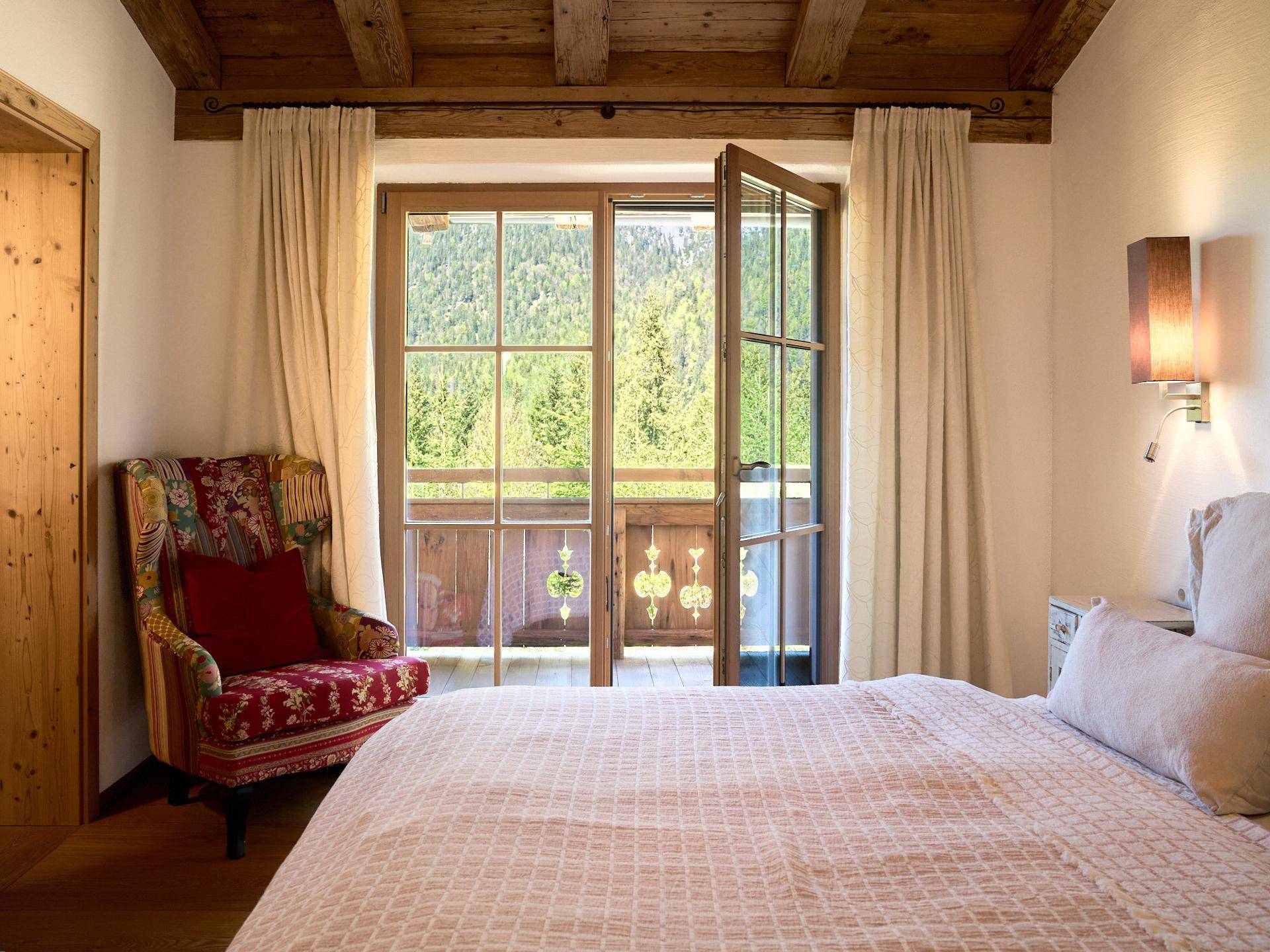 Gemütliches Schlafzimmer mit Holzbalkendecke, floralem Sessel und Balkonzugang.