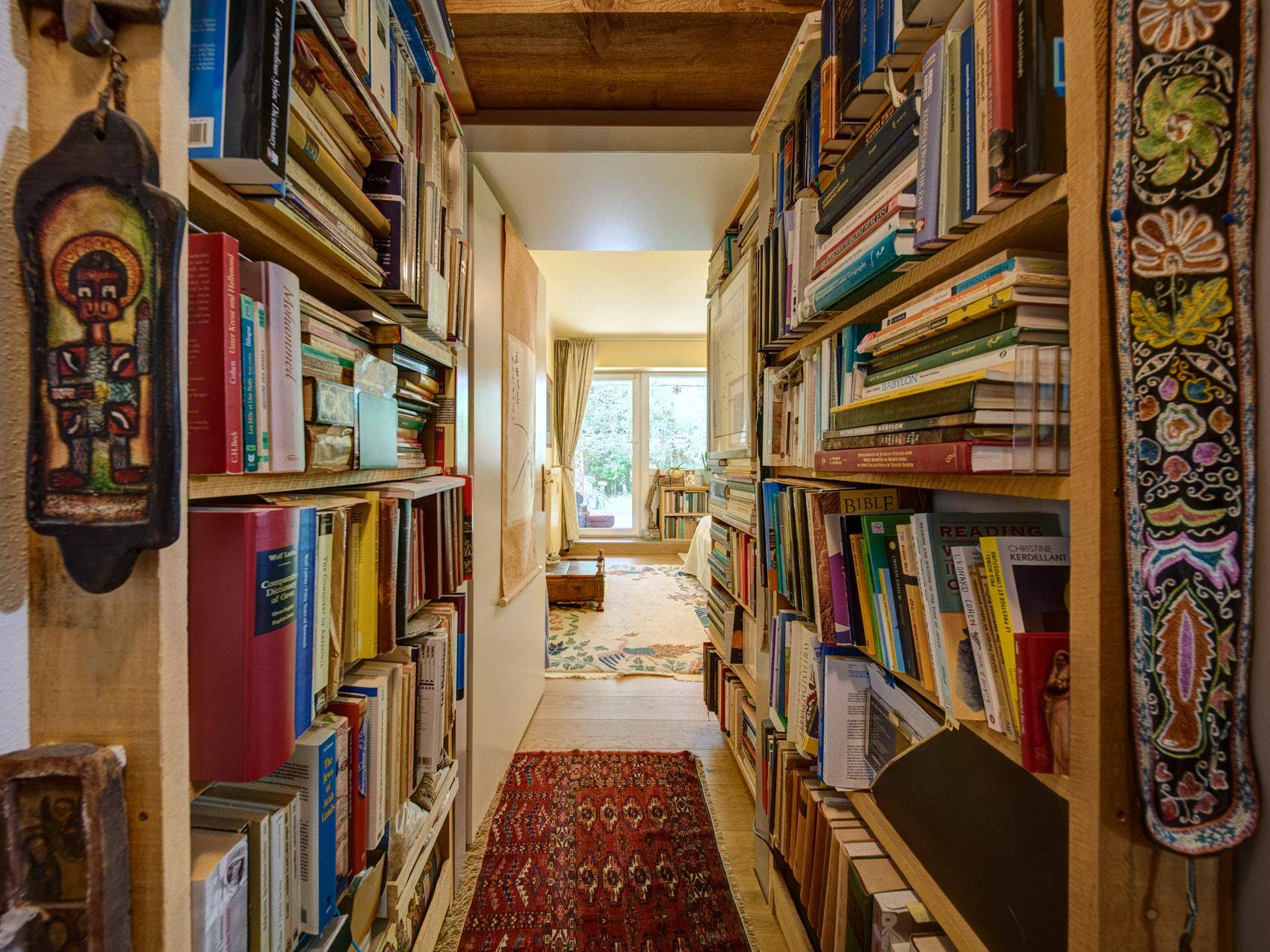 Schmaler Flur, gesäumt von hohen Bücherregalen, der zu einem hellen Raum mit Fenster führt.