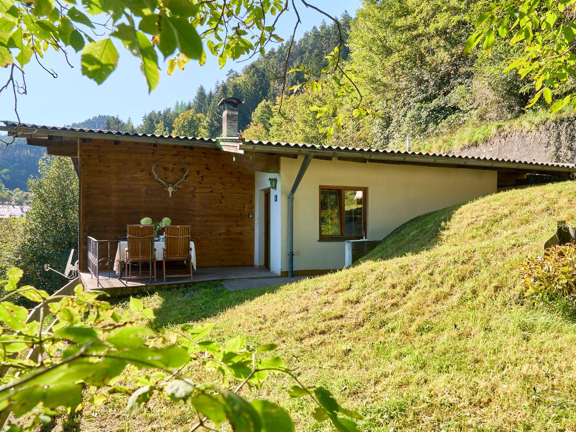Charmantes Haus mit Holzterrasse am Hang, umgeben von üppigem Grün und Hirschgeweih-Dekoration.