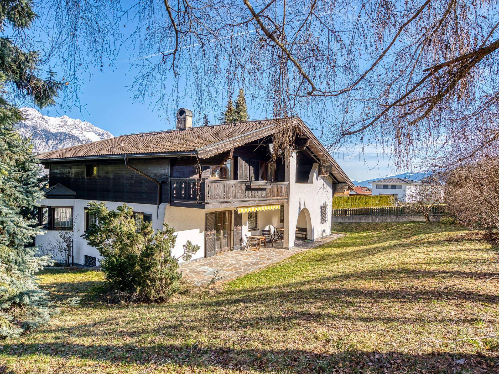 Traditionelles Haus mit Balkon, großem Garten und Bergblick unter blauem Himmel.