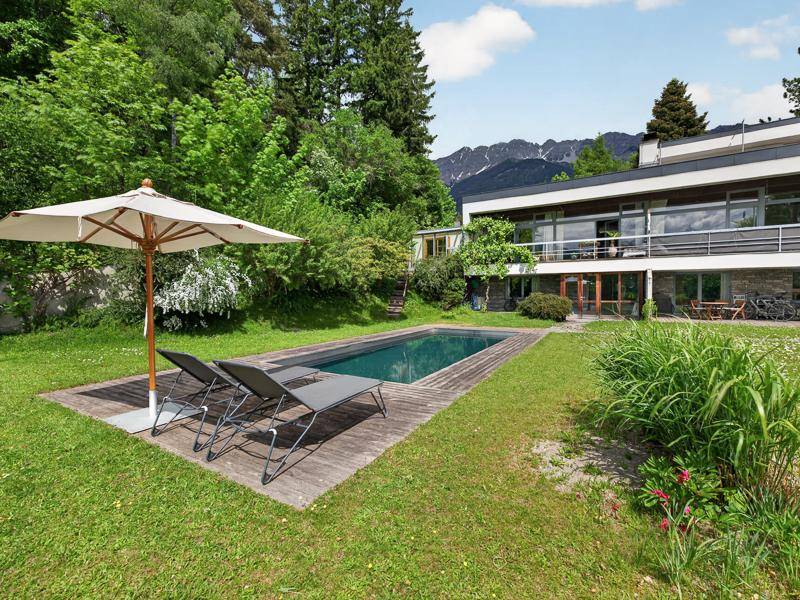 Garten mit rechteckigem Pool, Liegestühlen und Sonnenschirm, umgeben von Rasen und Bäumen.