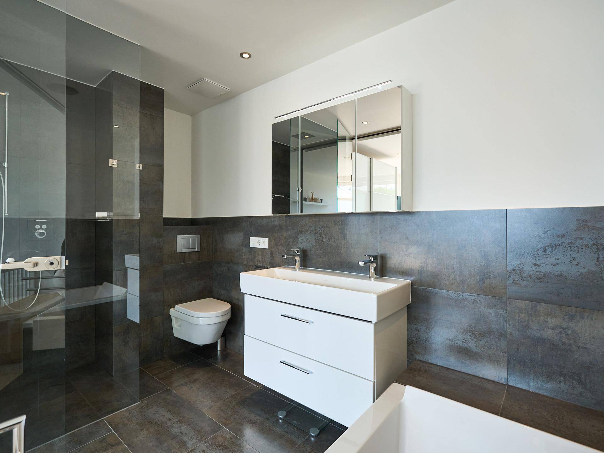 Modernes Badezimmer mit Doppelwaschbecken, großer Dusche und dunklen Fliesen.