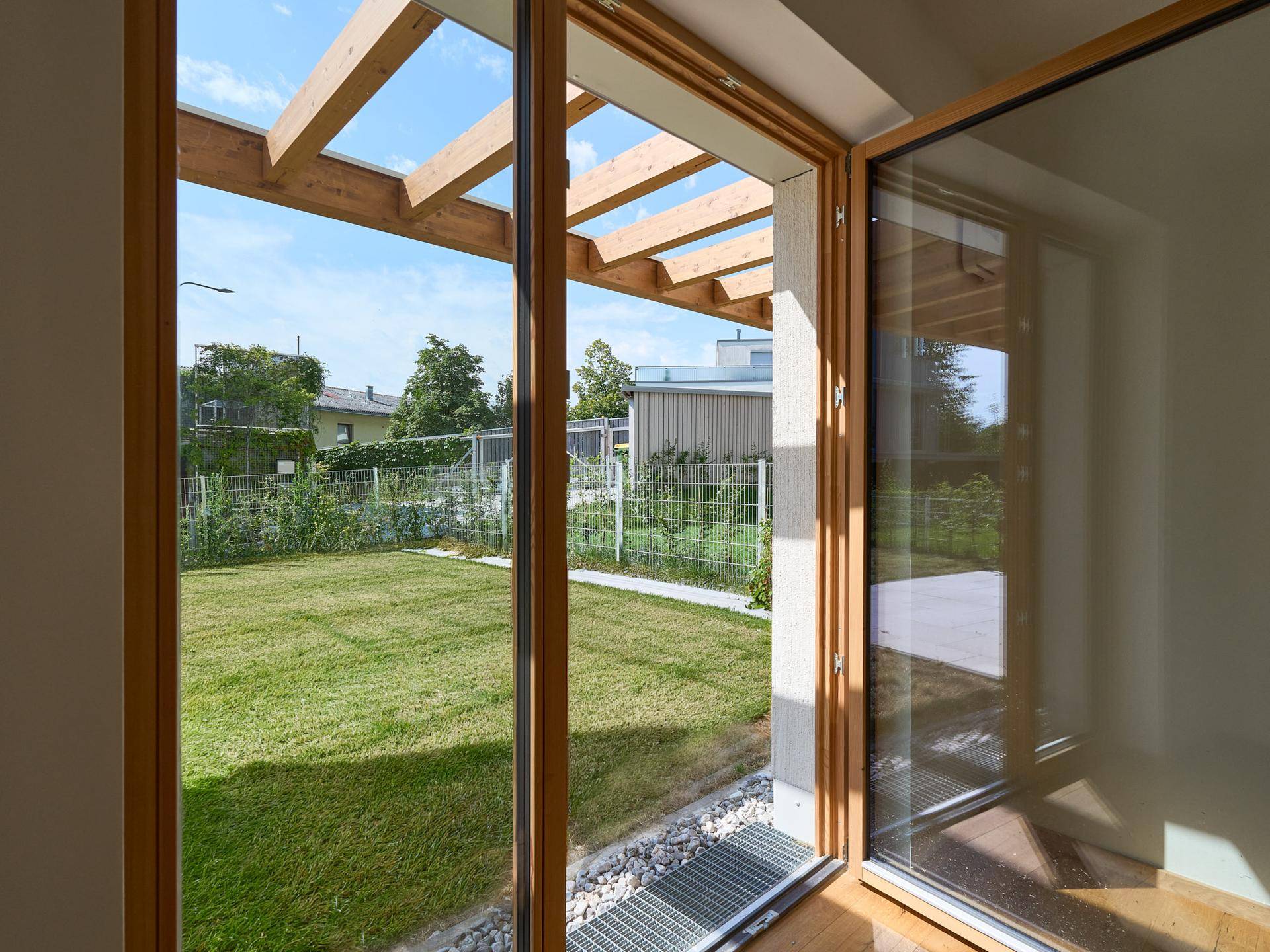 Blick aus dem Innenraum auf den grünen Garten mit Rasenfläche und Pergola.