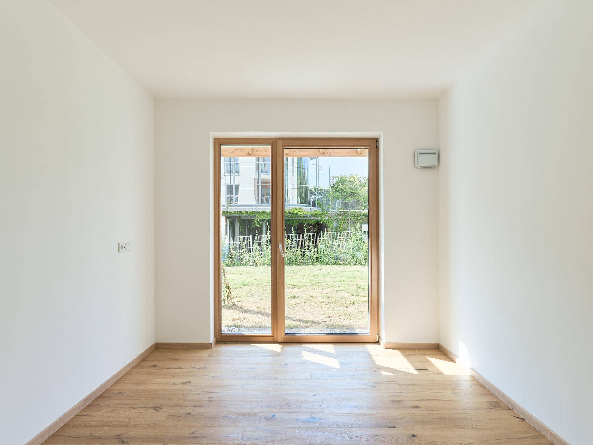 Helles Zimmer mit Holzboden und großen Fenstern, die den Blick in den Garten freigeben.