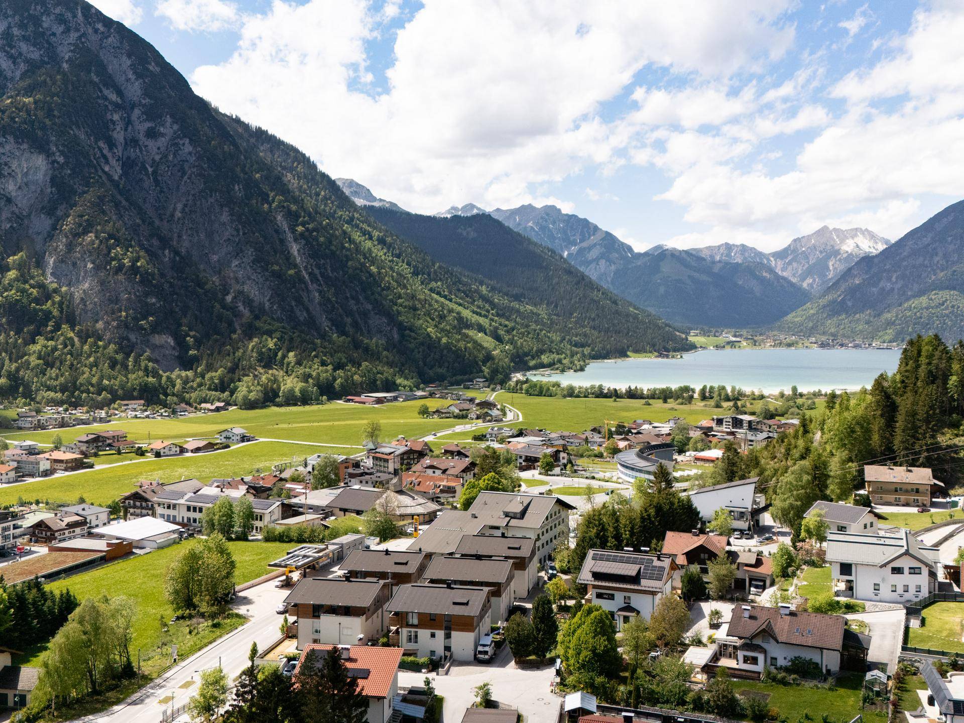Luftaufnahme eines Dorfes am Seeufer, umgeben von grünen Bergen unter einem bewölkten Himmel.
