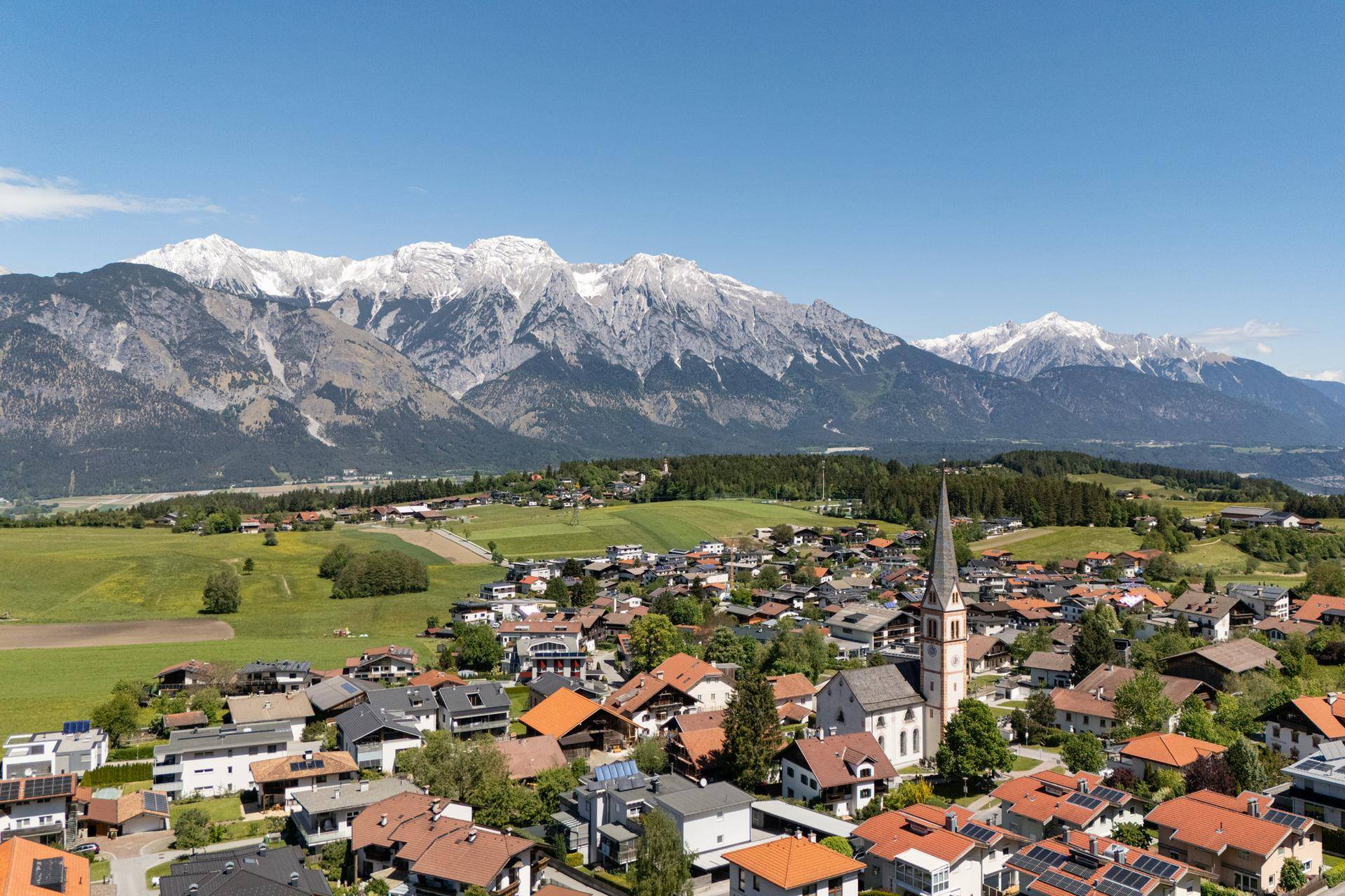 Panoramablick auf ein malerisches Dorf mit schneebedeckten Bergen im Hintergrund unter blauem Himmel.