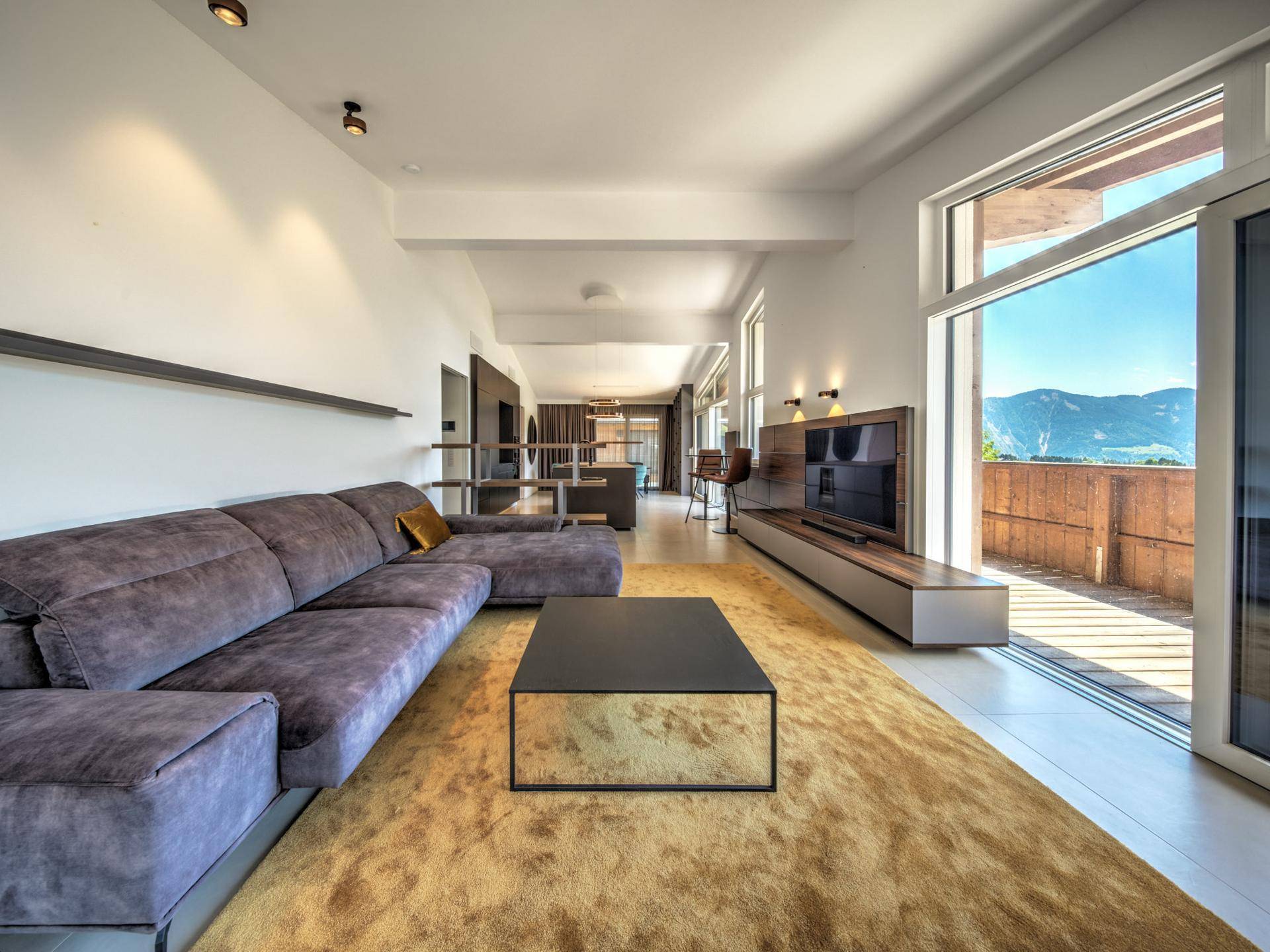 Geräumiges Wohnzimmer mit großem Sofa, Teppichboden und Zugang zum Balkon mit Bergblick.