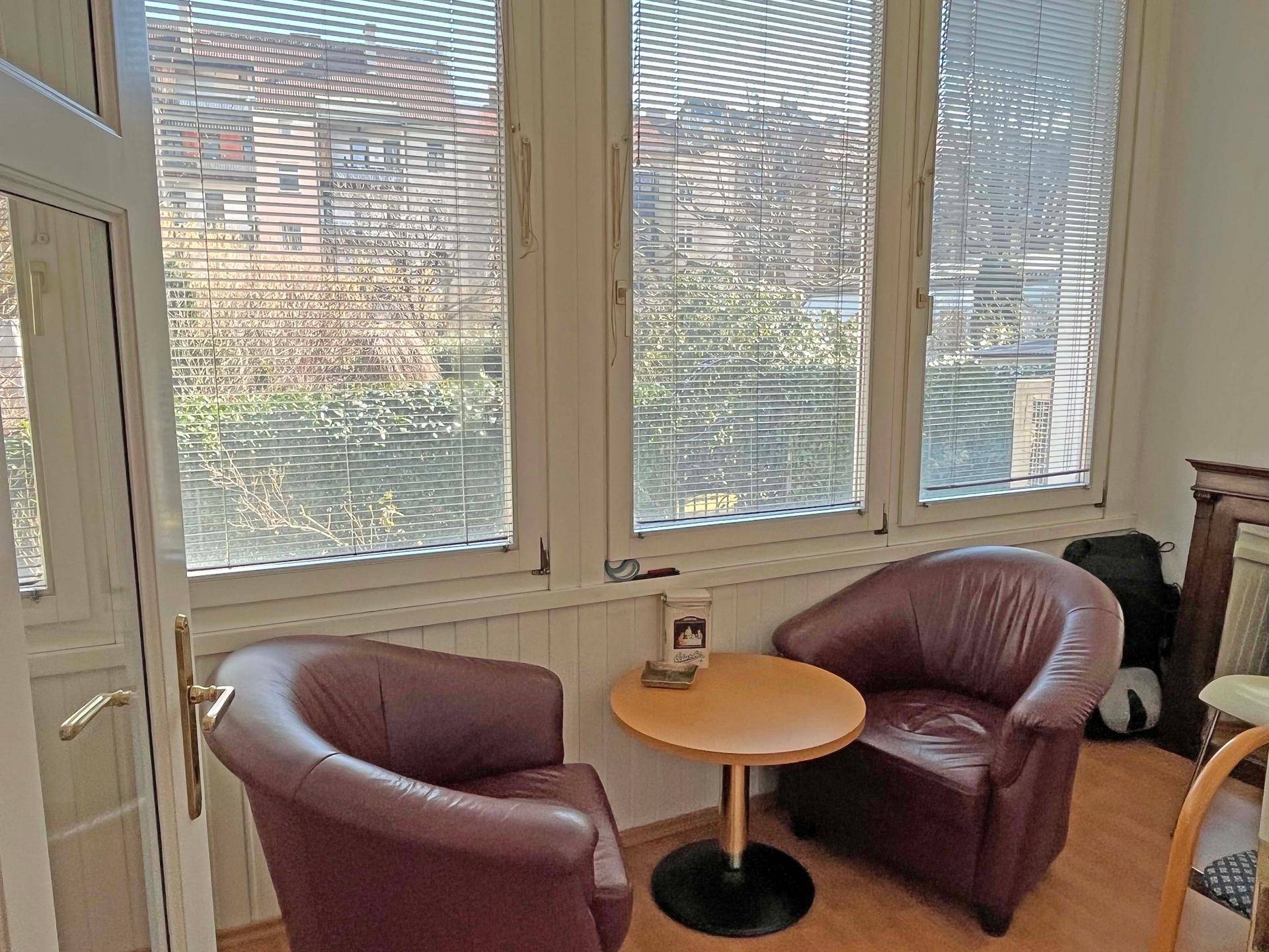 Gemütliche Sitzecke mit zwei Ledersesseln und kleinem Tisch vor großen Fenstern mit Jalousien.