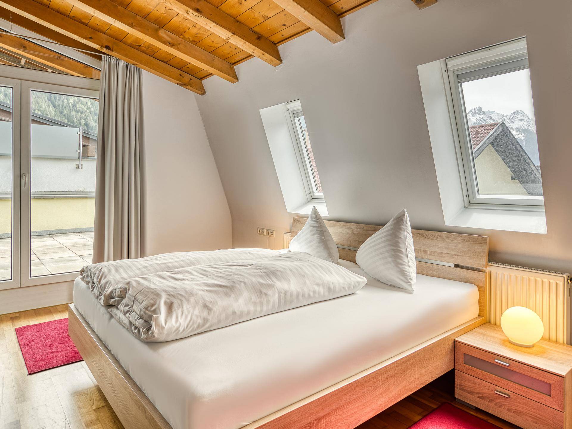 Helles Schlafzimmer im Dachgeschoss mit Holzbalken, Dachfenstern und Terrassenzugang.