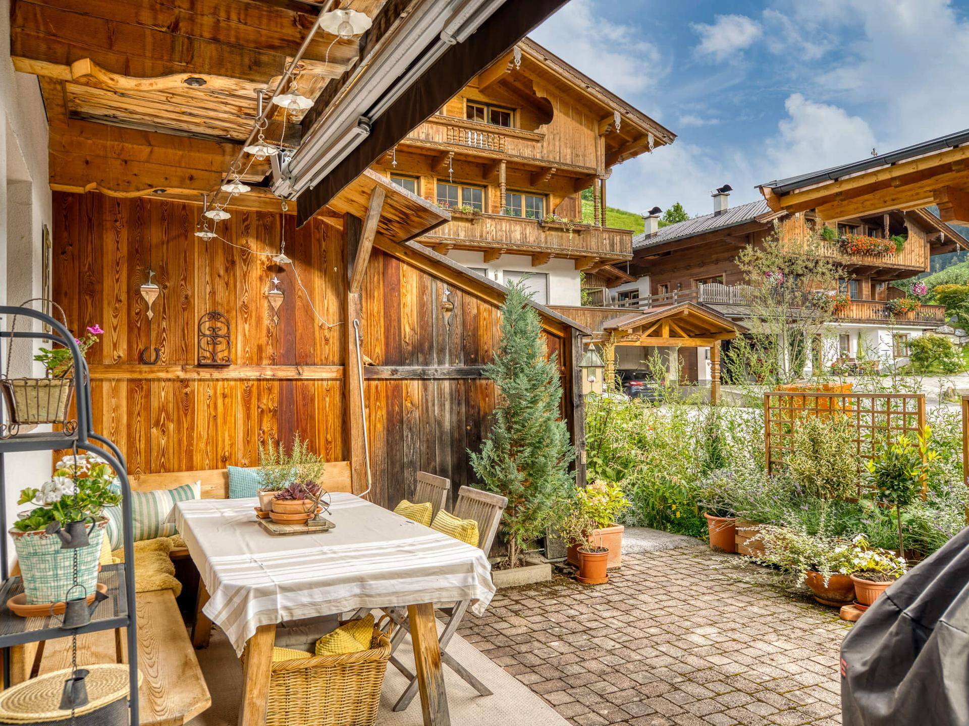 Überdachte Terrasse mit Esstisch und Sitzbänken, umgeben von Pflanzen und einem gepflasterten Bereich.