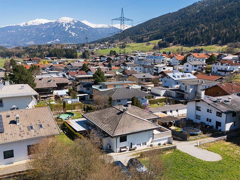 Luftaufnahme eines Wohngebiets mit vielen Häusern, umgeben von grünen Hügeln und majestätischen Bergen im Hintergrund.