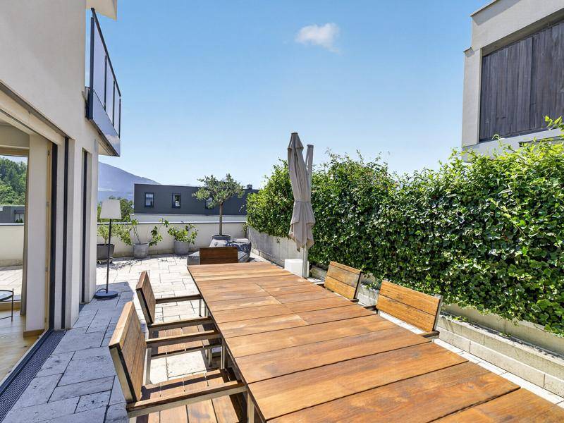 Geräumige Dachterrasse mit großem Holztisch, Sitzgelegenheiten und üppiger Bepflanzung unter blauem Himmel.