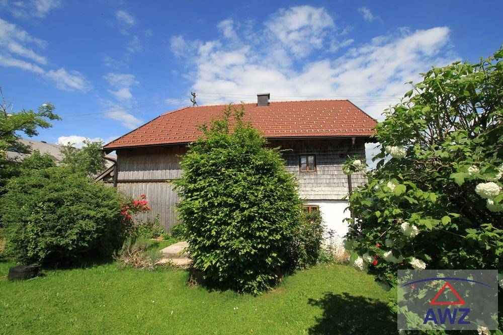 Traditionelles Holzhaus mit Satteldach, umgeben von üppigem Grün und blühenden Sträuchern unter blauem Himmel.