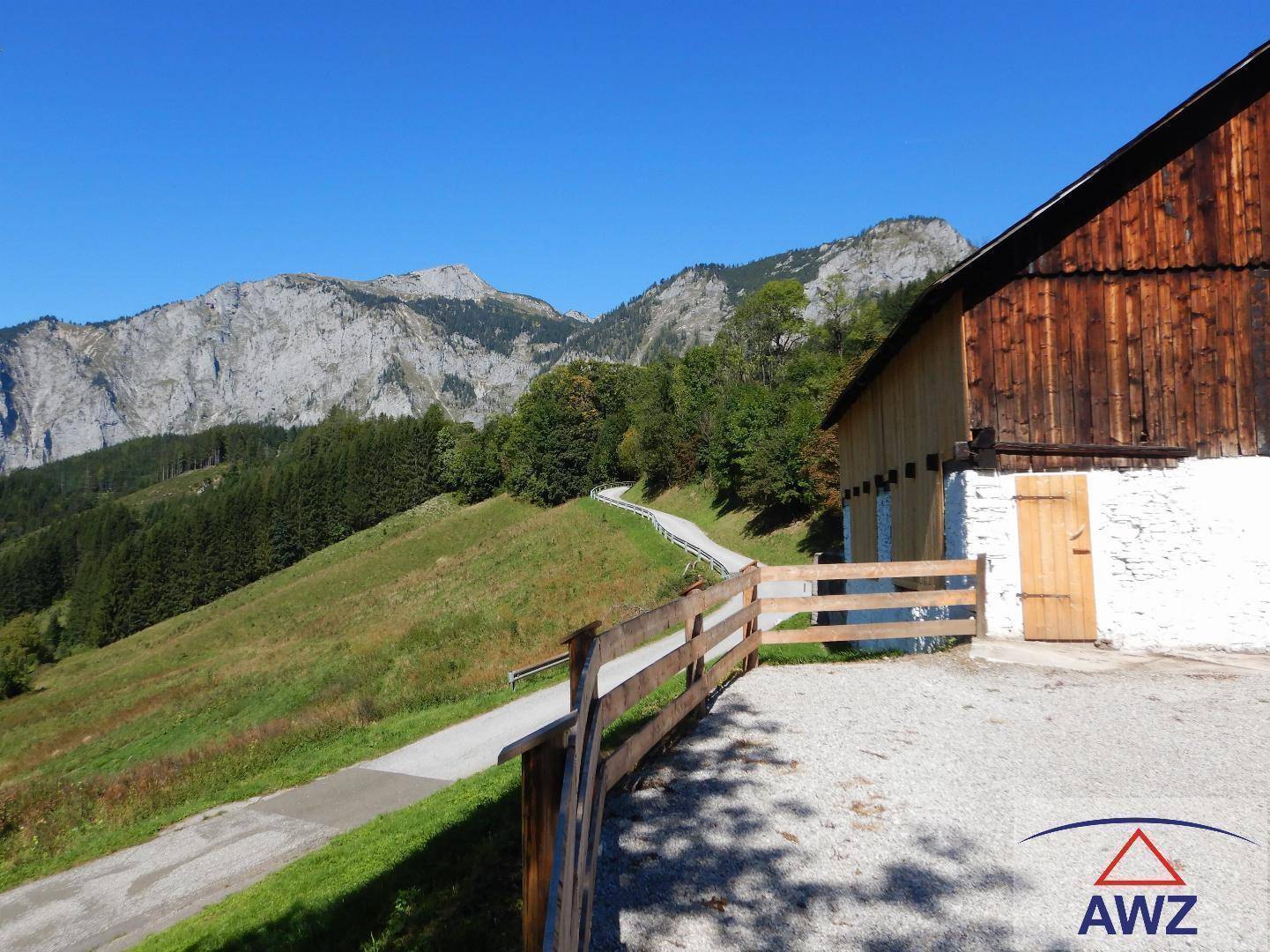 Rustikales Gebäude mit Holzelementen und weißem Sockel, umgeben von grünen Hügeln und beeindruckendem Bergblick.