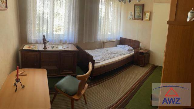 Schlafzimmer mit Einzelbett, Holzmöbeln und Teppichboden, hell durch zwei Fenster.