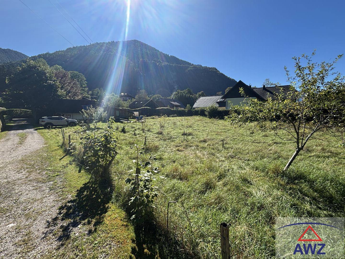 Sonniges Grundstück mit Grasfläche, Schotterweg und Häusern im Hintergrund, umgeben von Bergen.