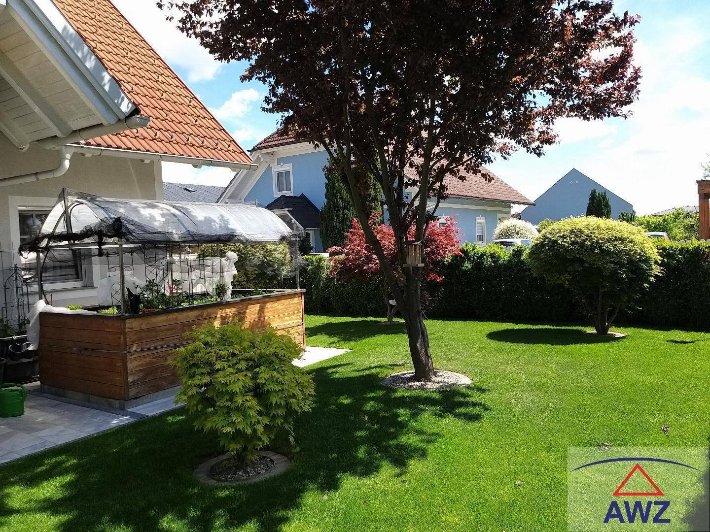 Gepflegter Garten mit Rasenfläche, Bäumen, Hecke und einem kleinen Gewächshaus unter blauem Himmel.