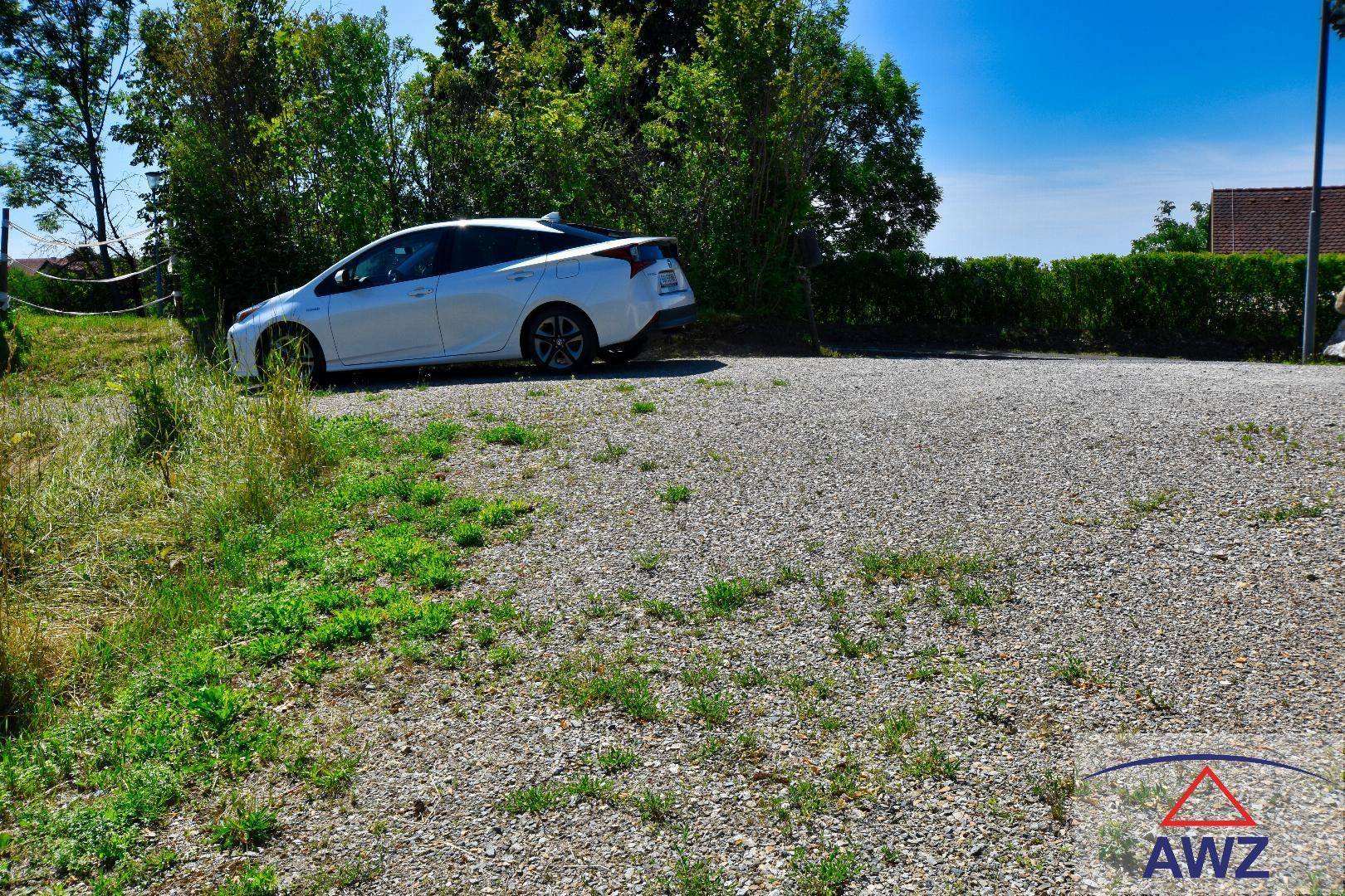 Schotterparkplatz mit einem weißen Auto, umgeben von grünen Büschen und Gras unter blauem Himmel.