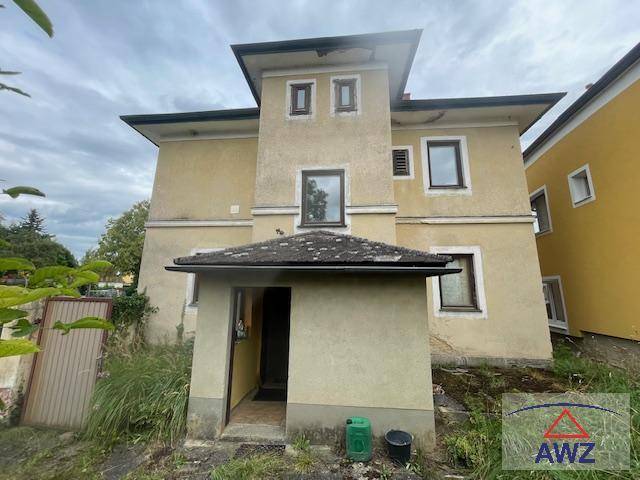 Außenansicht eines Hauses mit beiger Fassade, mehreren Fenstern und einem überdachten Eingangsbereich.