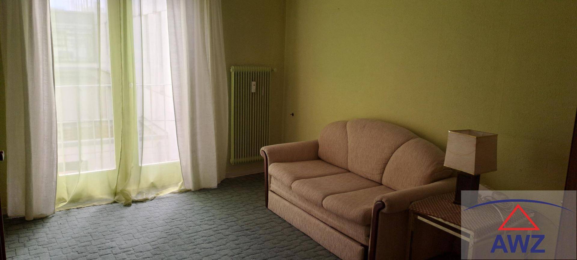 Gemütliches Zimmer mit beigem Sofa, grünem Teppich und großen Fenstern mit hellen Vorhängen.