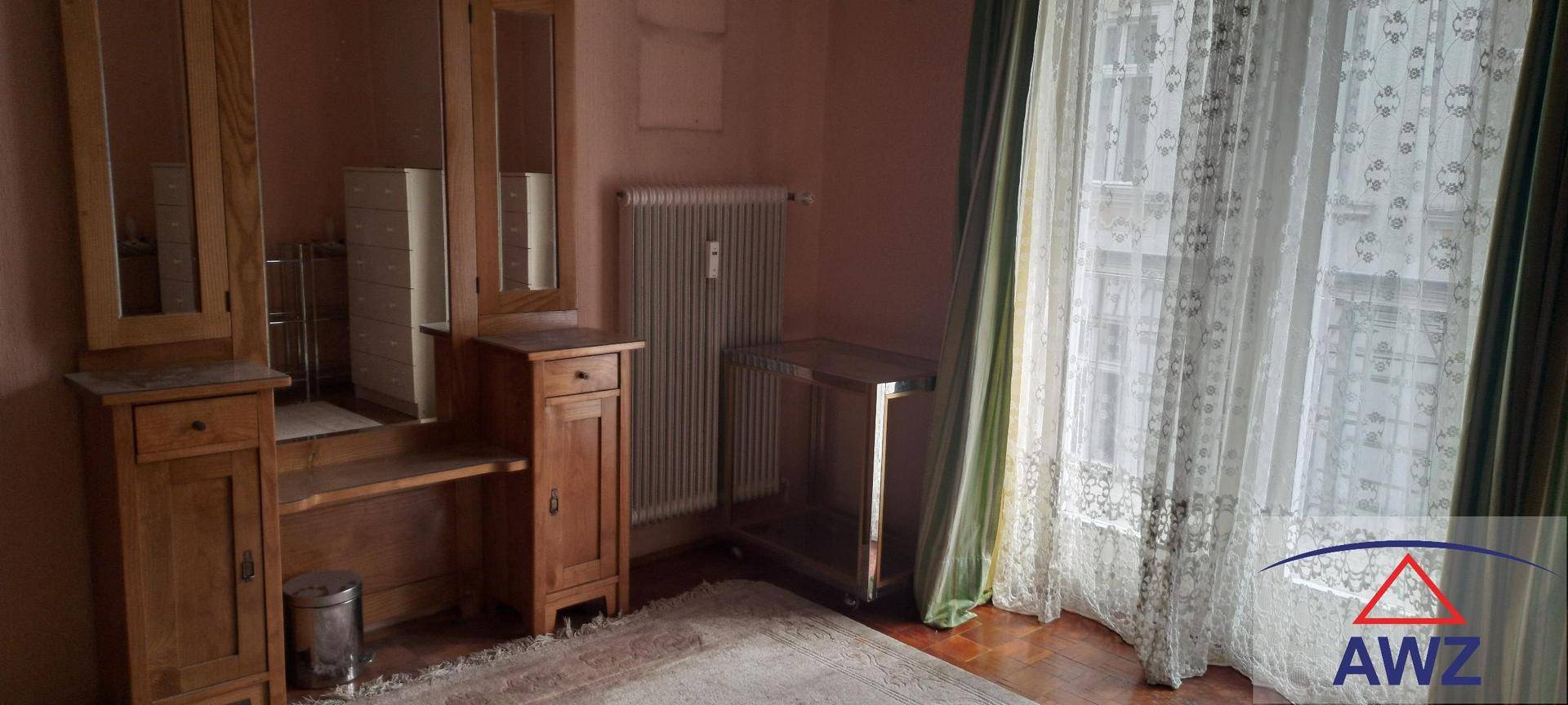 Zimmer mit Holzmöbeln, Heizkörper und einem Fenster mit gemusterten Vorhängen.