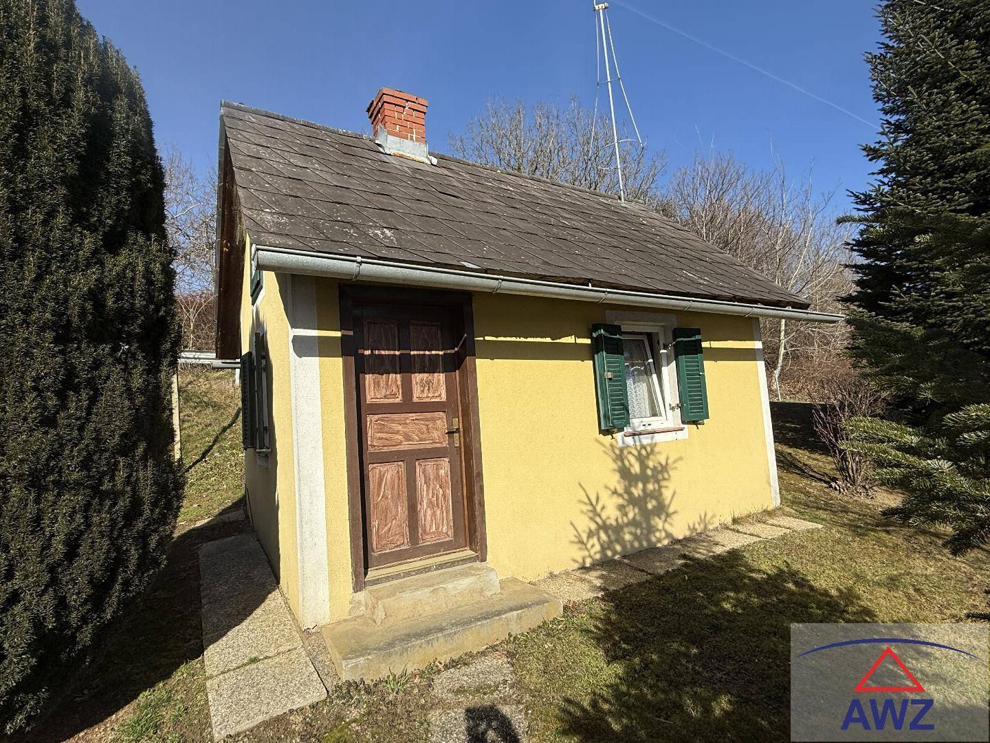 Kleines gelbes Gartenhaus mit Holztür und grünen Fensterläden, umgeben von Rasen und Bäumen.