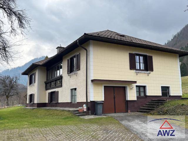 Seitenansicht eines Landhauses mit gelber Fassade, braunem Dach und Fensterläden, umgeben von Natur.