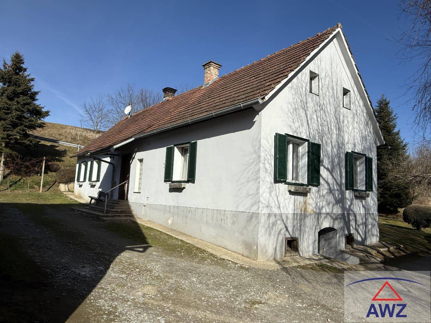 Charmantes Landhaus mit rotem Ziegeldach und grünen Fensterläden, umgeben von Bäumen und Natur.