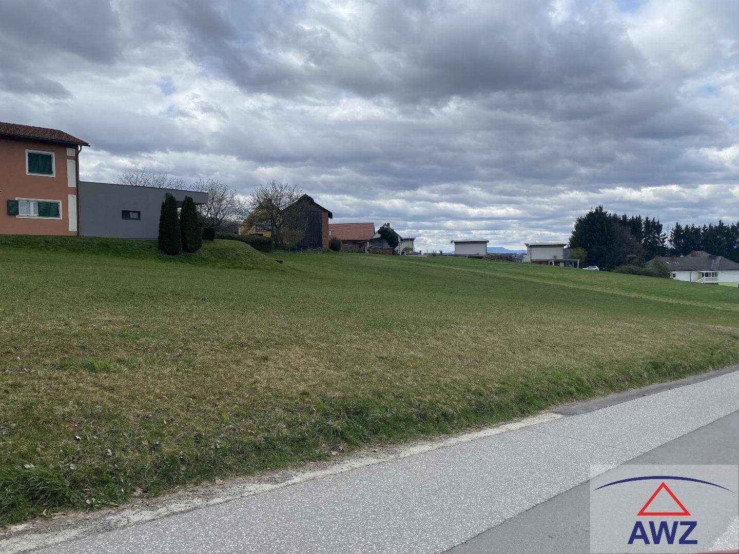 Grüne Hanglage neben einer asphaltierten Straße mit Wohngebäuden im Hintergrund unter bedecktem Himmel.