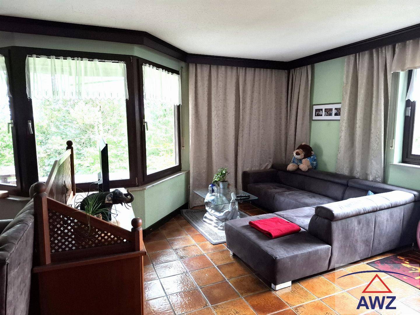 Gemütliches Wohnzimmer mit braunem Sofa, Terrakottafliesen und großen Fenstern mit Blick ins Grüne.