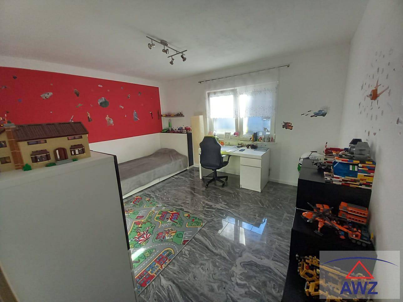 Kinderzimmer mit roter Akzentwand, Schreibtisch, Bett und Spielzeug auf glänzendem Fliesenboden.