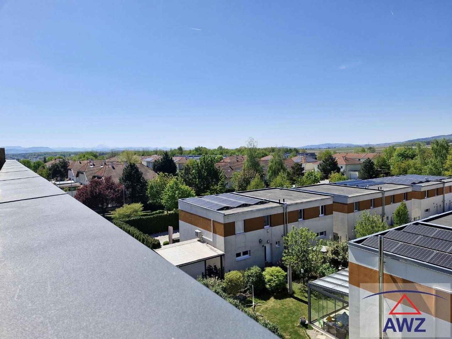 Weitblick über eine Wohnsiedlung mit viel Grün und fernen Bergen unter blauem Himmel.