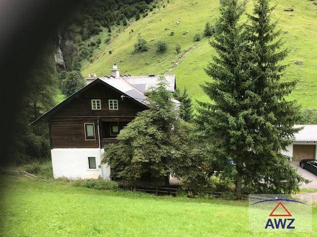 Traditionelles Holzhaus in alpiner Landschaft mit grünen Wiesen und hohen Bäumen.