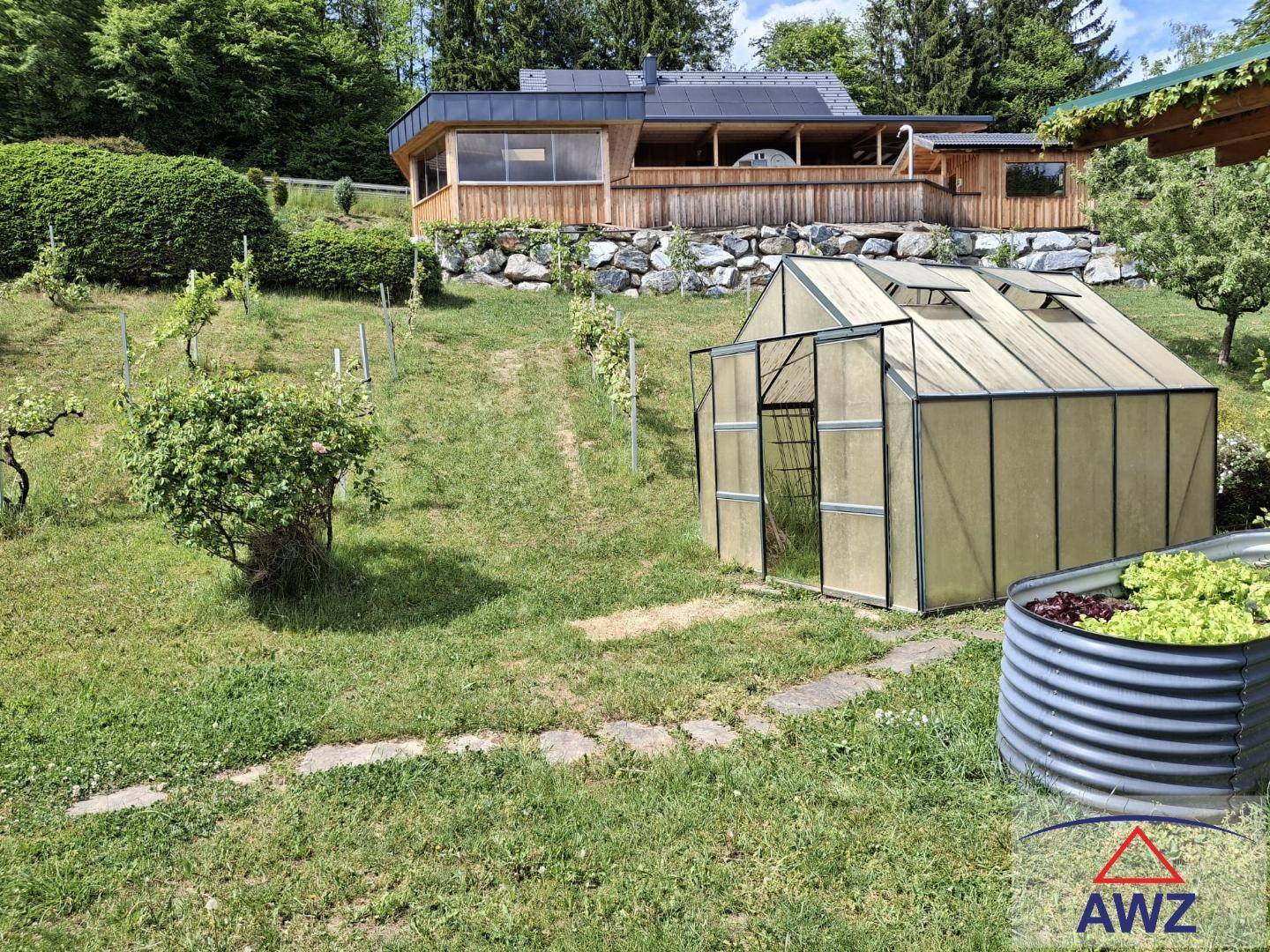 Sonniger Gartenbereich mit Gewächshaus, Hochbeet und Weinreben, im Hintergrund ein Holzhaus mit Terrasse.