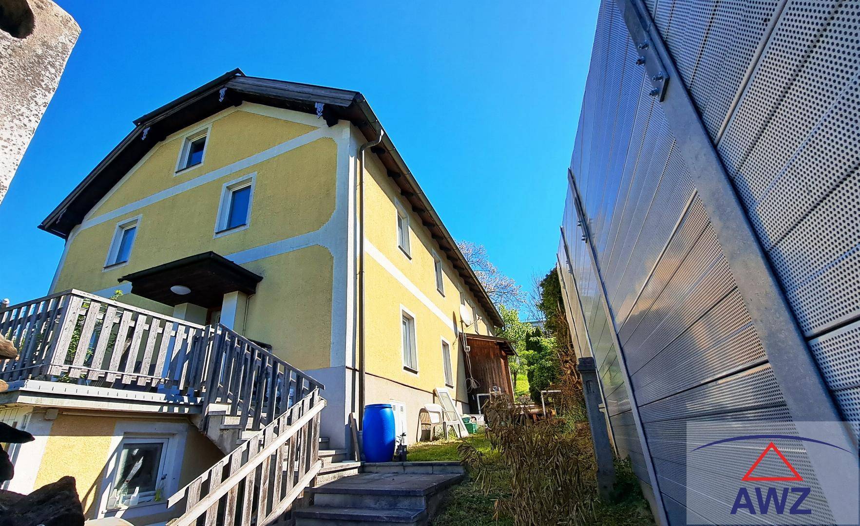 Ansicht der Hausrückseite mit Außentreppe und angrenzender moderner Metallwand unter strahlendem Himmel.