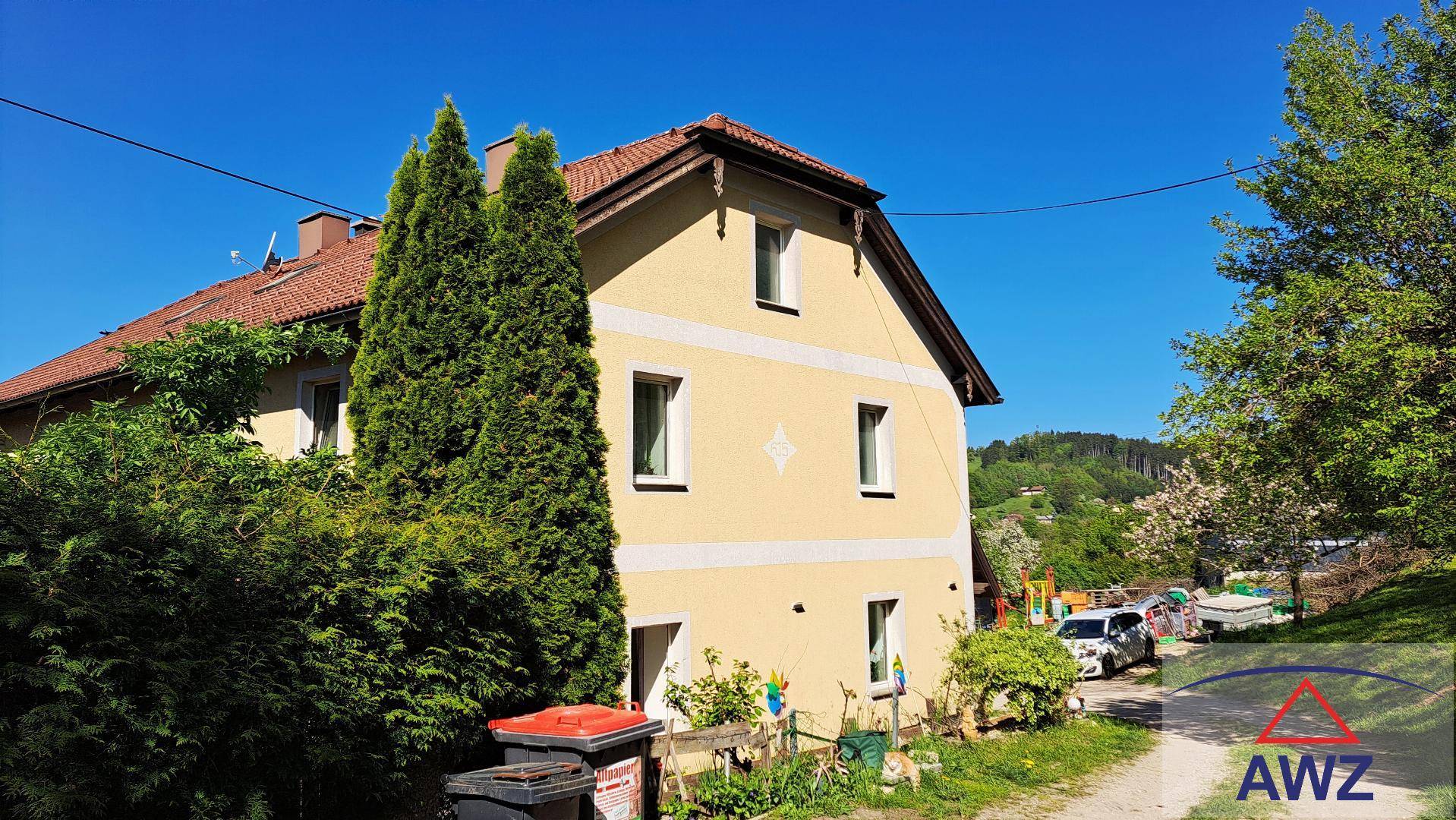 Seitenansicht des Hauses mit gelber Fassade, roten Dachziegeln und hohen Nadelbäumen im Vordergrund.