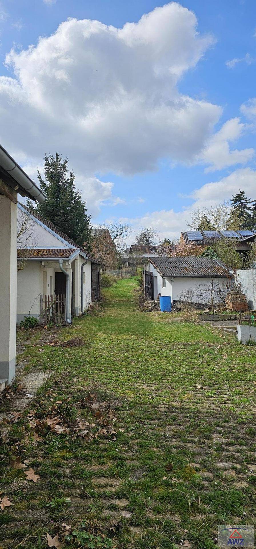 Langer, grasbewachsener Bereich zwischen mehreren kleinen Nebengebäuden unter einem bewölkten Himmel.