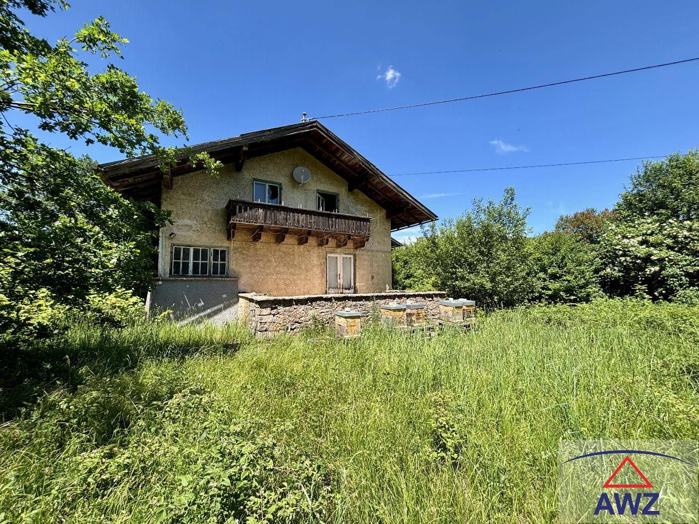 Älteres Wohnhaus mit Balkon und Terrasse, umgeben von üppiger Vegetation und Wiesen.