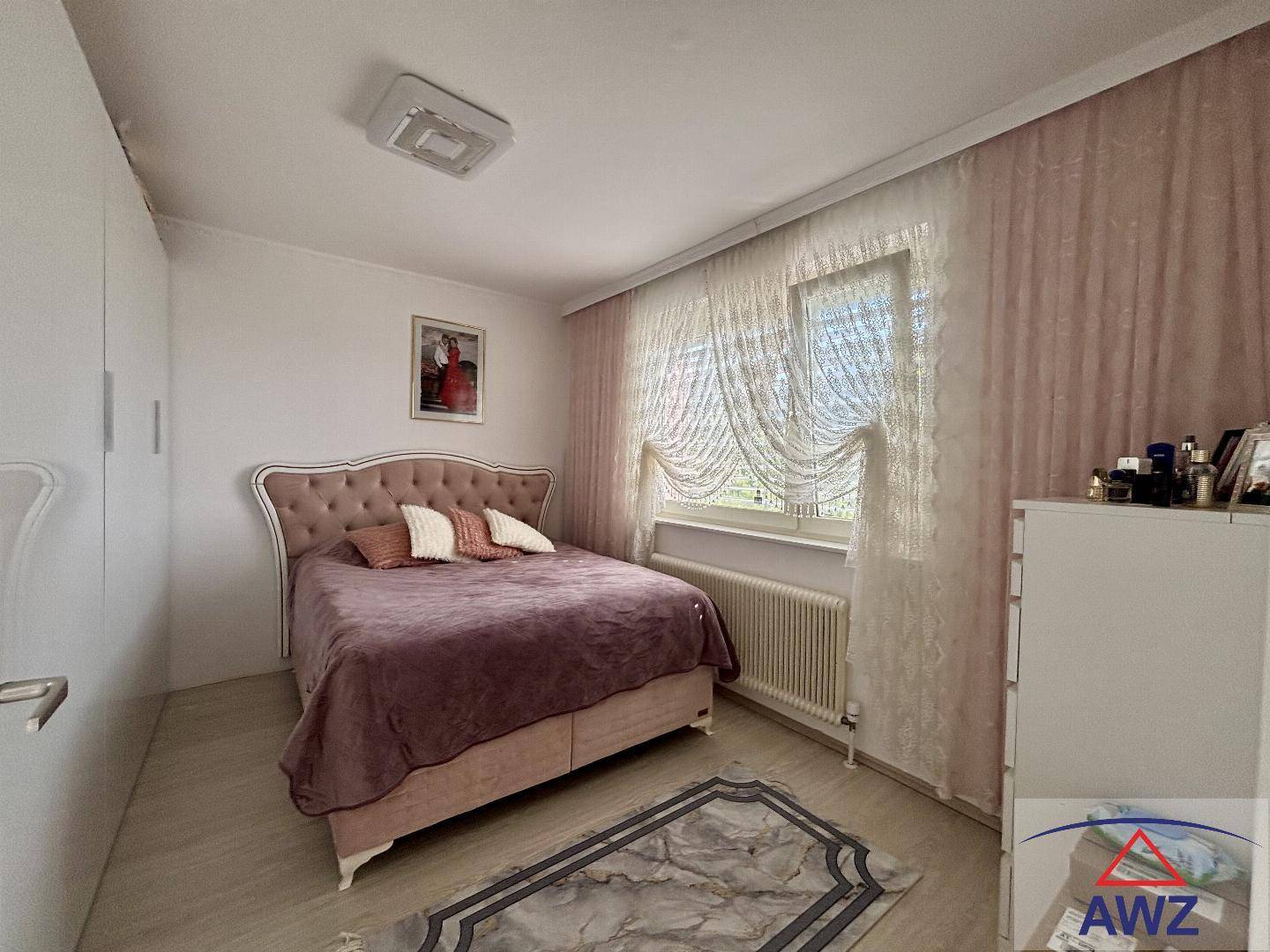 Gemütliches Schlafzimmer mit großem Bett, hellem Laminatboden und Fenster mit Vorhängen.