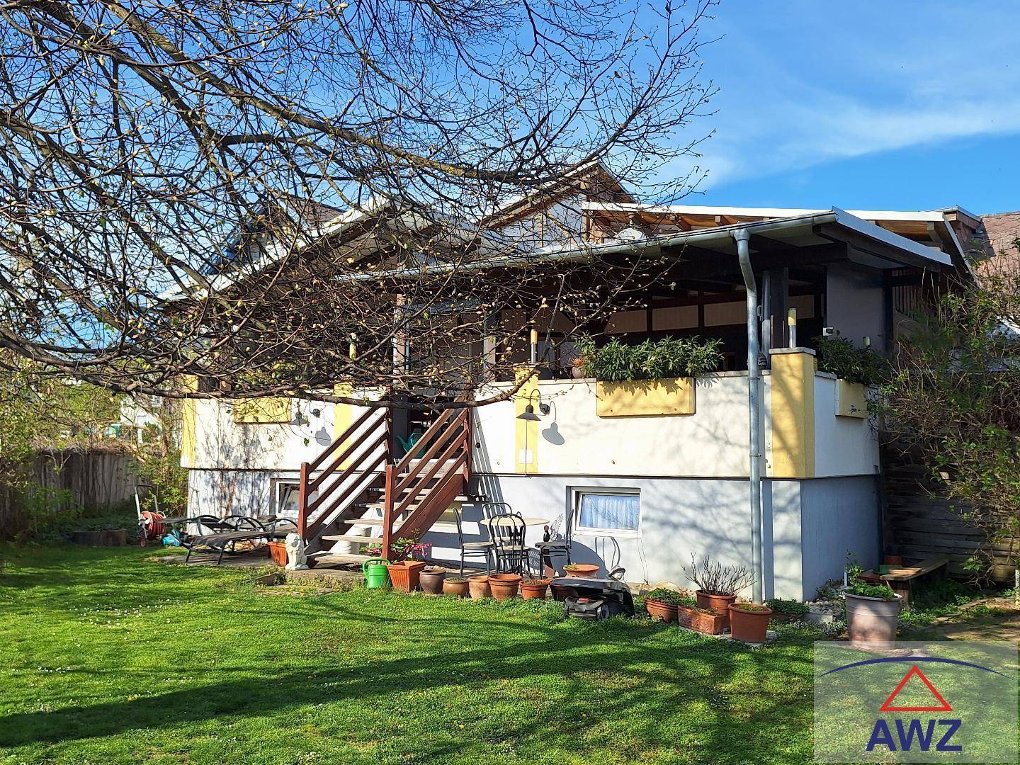Hausfassade mit überdachter Terrasse und großem Gartenbereich unter blauem Himmel.