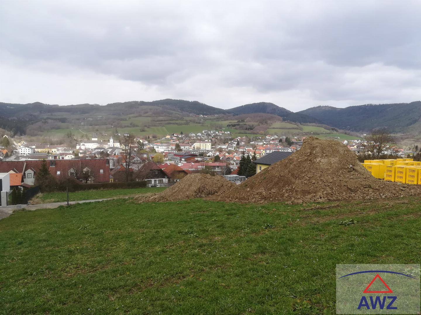 Blick auf ein Baugrundstück mit Erdhügeln und einer Ortschaft im Hintergrund unter bewölktem Himmel.