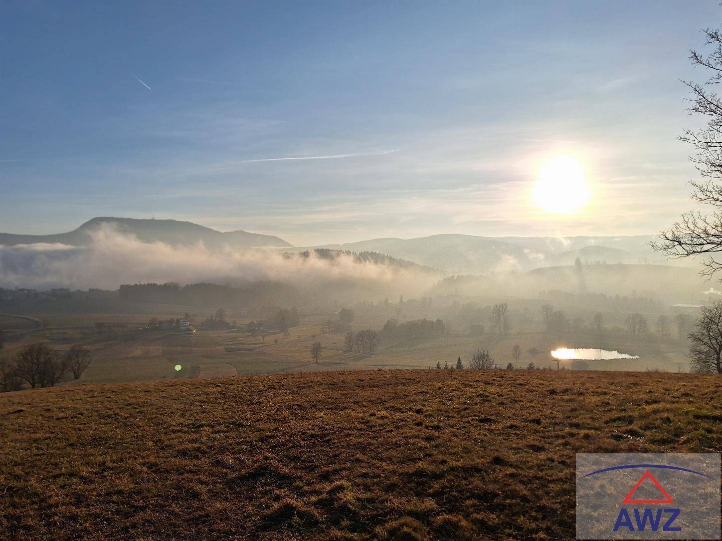 Panoramablick über eine neblige Landschaft mit Bergen, einem kleinen See und der aufgehenden Sonne.
