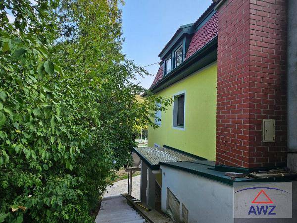 Ansicht der Hausseite mit gelber Fassade, rotem Dach und einem markanten Backsteinschornstein.
