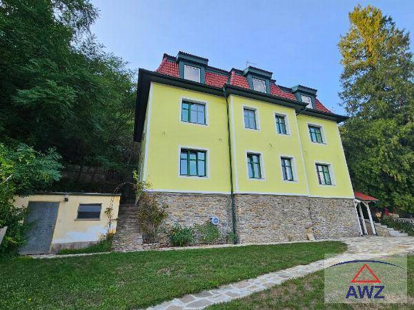 Gesamtansicht des Hauses mit gelber Fassade, rotem Mansarddach und Steinsockel, umgeben von Bäumen.