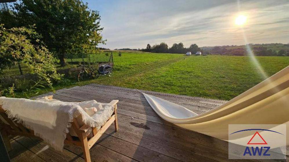 Sonnige Holzterrasse mit Hängematte und Sitzbank, umgeben von weiten grünen Feldern.