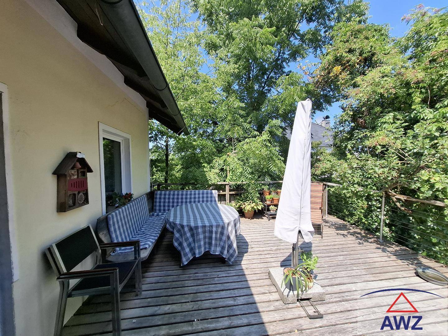 Großzügige Holzterrasse mit Sitzgelegenheiten und Sonnenschirm, umgeben von Bäumen.