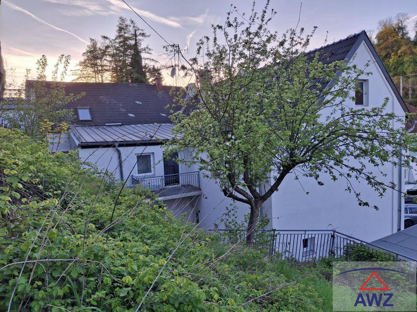 Das weiße Gebäude eingebettet in eine grüne, hügelige Landschaft mit einem blühenden Baum im Vordergrund.