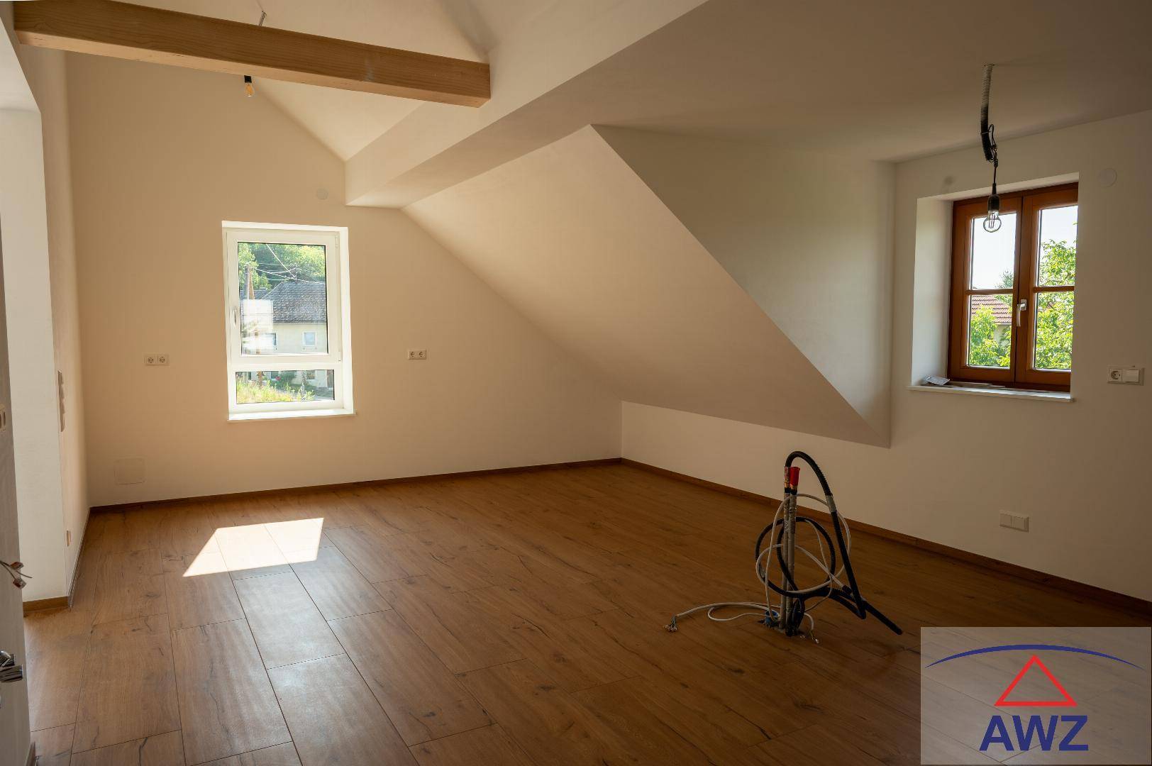 Lichtdurchfluteter Raum mit Dachschrägen, Holzbalken und zwei Fenstern, die viel Helligkeit spenden.