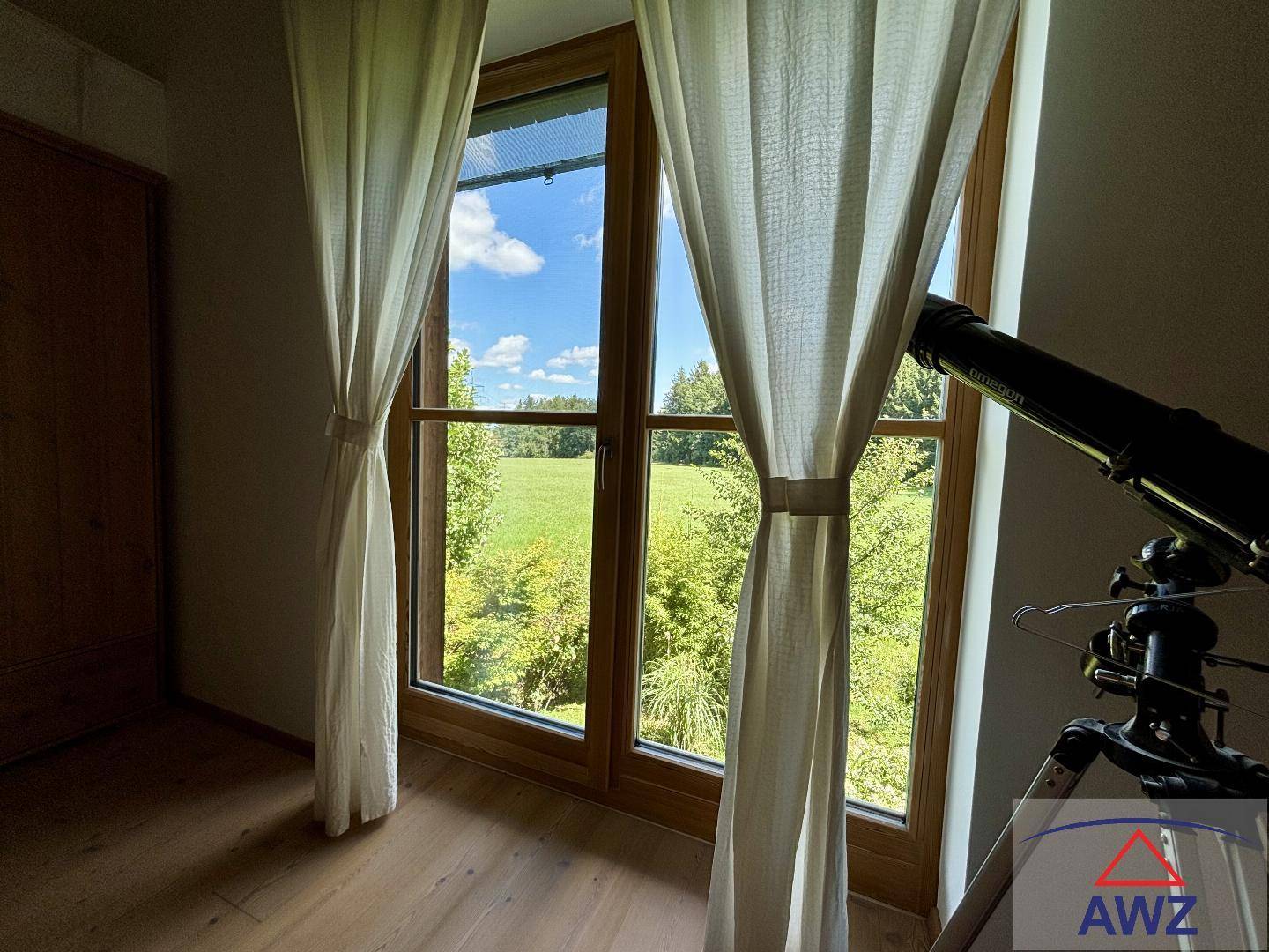 Fenster mit Vorhängen und einem Teleskop, das einen weiten Blick auf grüne Felder und Wälder bietet.