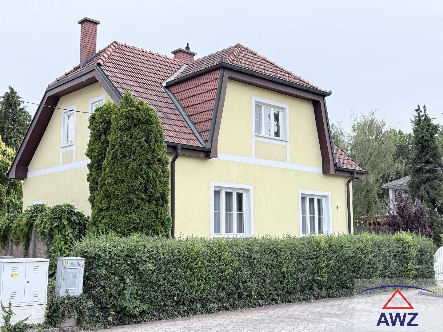 Ansicht des gelben Einfamilienhauses mit Mansarddach und grüner Hecke im Vorgartenbereich.